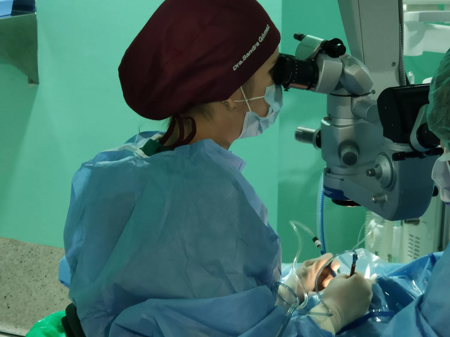 Cirujana usando un microscopio durante una operación en un quirófano, con equipo médico y personal en un ambiente clínico.